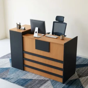 A modern L-shaped reception desk featuring a dual-tone design with charcoal gray side panels and natural oak wood front faces, incorporating an elevated transaction counter, an inner workspace desk, and an attached vertical storage tower with one lockable drawer and one lower cabinet door.